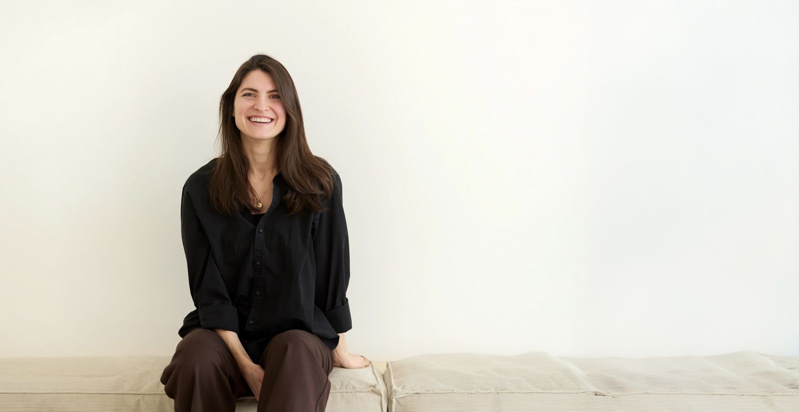 Nelly Dommel, Gr&uuml;nderin von Studio Nedo, sitzt lachend auf einer Bank vor heller Wand
